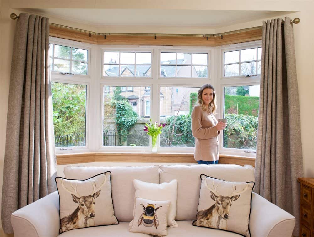 Model photographed with a cup of coffee in a living room with large bay window. Cream sofa and curtains.